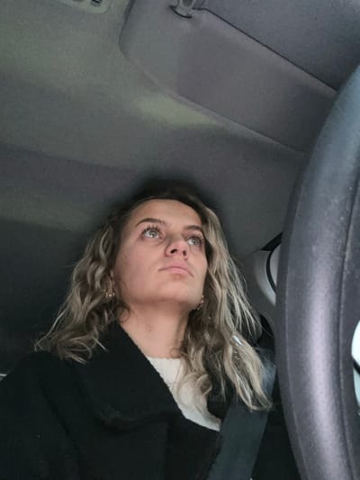 A blonde woman with light eyes and curly hair lies in a car, looking upwards. The car's steering wheel is above her head. She is wearing a dark jacket over a white shirt or sweater. The car's interior, including the ceiling and side, are visible and are gray.