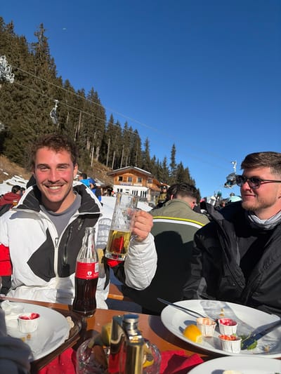 The image depicts an outdoor dining scene at a ski resort on a clear, sunny day. The location is identifiable as the Sportalm Hütte in Schwendau, Austria, with the hut's name clearly visible on a wooden building in the background.

In the foreground, two adult males are seated at a wooden table covered with a red tablecloth. The male on the left, with short dark curly hair and a mustache, is smiling directly at the camera. He is wearing a white, black, and red ski jacket over a grey top. His right hand holds a large glass mug containing golden liquid, consistent with beer, and his left hand rests on a classic glass Coca-Cola bottle positioned on the table. The male on the right, partially obscured, wears dark sunglasses and a black ski jacket with a light-colored neck gaiter, looking towards the right of the frame.

The table holds several white plates, some containing remnants of food, including small white paper cups with red or green contents, and lemon wedges. A metallic, silver-toned object, possibly a cocktail shaker, is also present on the table.

In the mid-ground, beyond the table, other patrons in winter attire are visible around and in front of the Sportalm Hütte. The ground is covered with snow.

The background features a snow-covered mountain slope densely forested with pine trees. A ski lift system with visible chairs extends upwards along the mountain. The sky is bright and clear blue, with a small, light-colored paraglider visible high in the upper-left quadrant.