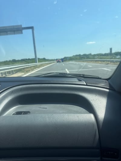 A view from inside a vehicle driving on a highway near Zagore, Bulgaria. The highway is multi-lane with a median and roadside vegetation. There are other vehicles visible in the distance. A road sign is present on the left side of the highway. The interior of the car is visible, including the dashboard and door panel.