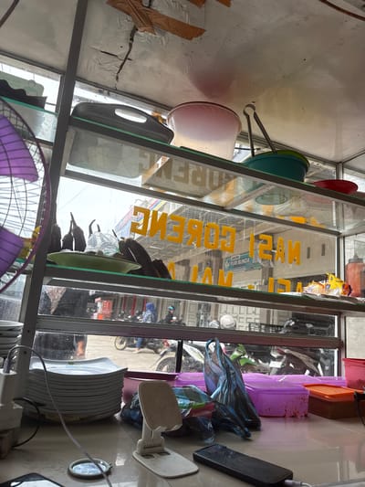 The image shows a street food stall in Meunasahmanyet, Indonesia. The stall is made of metal and glass, and it is filled with various items for sale. On the top shelf, there are some plastic containers and a pink bowl. On the second shelf, there are some eggplants in a green bowl, and a plastic bag. On the third shelf, there are some bags of snacks and an orange container. On the bottom shelf, there are some plates, a phone charger, and a smartphone. The background shows a street with several motorcycles and people. The overall impression is that of a busy and colorful street food stall.