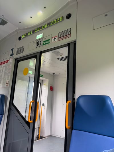 The image captures the interior of a passenger train car. In the foreground, a blue seat is visible on the right. A closed sliding door with orange handles is on the left, revealing a glimpse of the interior beyond. Above the door, a digital display shows the date and time: "07:01 23.06.2026". Adjacent to the time display are signs indicating "АВАРИЙНЫЙ ВЫХОД" (Emergency Exit) and "01 ВЫХОД" (01 Exit). Further to the right, there are smaller signs, one with a camera icon and text that appears to be a warning about video surveillance, and another red sign with "НЕ КУРИТЬ!" (Do not smoke!). To the left of the door, a sign with the number "1" and the word "ВАГОН" (Car) is prominently displayed. The overall environment suggests a clean and modern train interior. The prompt mentions the city of Баграш-Бигра, Russia, but this information is not visually verifiable from the image itself.
