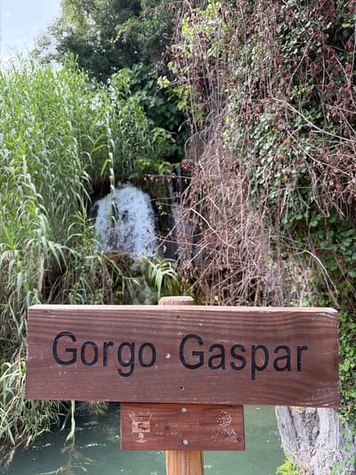 The image shows a wooden sign with the text "Gorgo Gaspar" carved into it, in the town of Anna, Spain. Below this sign, there is another smaller wooden sign with a crest and the text "Ayuntamiento de Anna" and "ANNA. MANANTIAL DE LEYENDA". The signs are placed in front of a natural landscape featuring a waterfall, lush green vegetation, and a body of water. The overall scene depicts an outdoor setting, likely a tourist or recreational area.