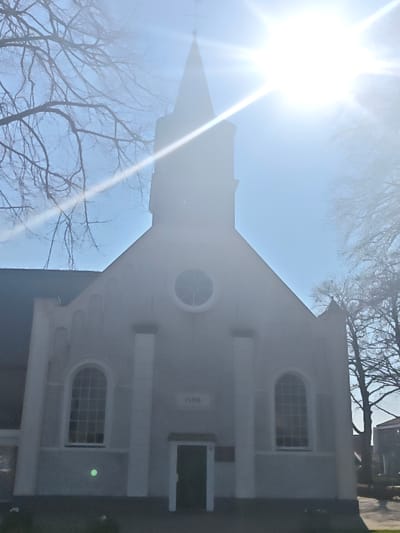 The image depicts the front facade of a church building located in Wierden, Netherlands, under bright, direct sunlight. The building features a light-colored, likely stucco, exterior with a gabled roof. A prominent dark entrance door is centered at ground level, beneath a small white canopy. Flanking the entrance are two tall, arched windows with multi-pane glazing. Above the entrance, centered on the gable, is a circular window, also with multi-pane glazing. A date, "1909," is visibly inscribed or carved into the facade directly above the entrance and below the circular window. Vertical, light-colored pilaster-like architectural elements adorn the facade. Rising from the roofline is a dark, silhouetted steeple with a pointed spire, topped by a cross. The sky is clear blue, with a strong sunburst in the upper right quadrant casting significant lens flare and backlighting the scene. Bare deciduous tree branches are visible on both the left and right sides of the frame, with more prominent branches in the foreground on the left. The ground in the immediate foreground is dark. Faint outlines of other structures are visible in the distant background to the right. No individuals or discernible actions are present.