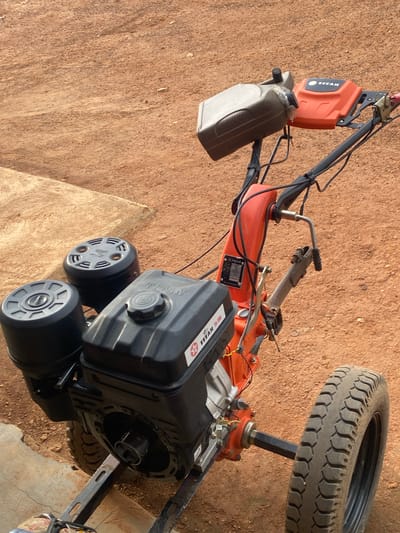 An outdoor, eye-level, medium shot captures a walk-behind tiller in Jere South, Nigeria. The machine, predominantly orange and black, rests on a reddish-brown dirt surface, suggestive of a rural or unpaved urban environment. The tiller's engine is black and features the "TITAN" brand name. Two black cylindrical air filters are attached to the engine. Above the engine, an orange cover with "TITAN" branding is visible, alongside a gray plastic container, possibly for fuel or oil, secured with tape. The tiller has a single tire on the right side, showing a worn tread pattern. The background consists of more of the same reddish-brown dirt. The lighting suggests it is daytime. No people are present in the frame.