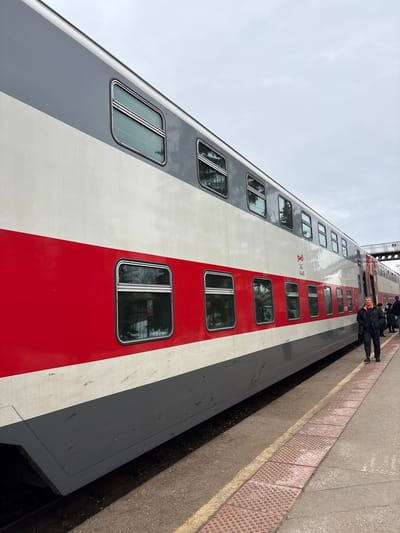 A bi-level passenger train is stopped at a concrete platform under an overcast sky in Dimitrovgrad, Russia. The train car is striped horizontally in grey (top), white (upper middle), red (lower middle), and dark grey (bottom). The "РЖД" logo and the text "065 144-40" are visible on the white section. Multiple rectangular windows with rounded corners are present on both levels, reflecting the environment. The platform features a reddish-brown tactile paving strip and a yellow safety line. In the immediate foreground, a man, wearing a dark jacket with an orange lining and a dark cap, walks on the platform parallel to the train, facing left. Further down the platform to the right, other individuals are visible near an open train door, consistent with passenger activity. A metal pedestrian overpass is partially visible in the distant background above the train.