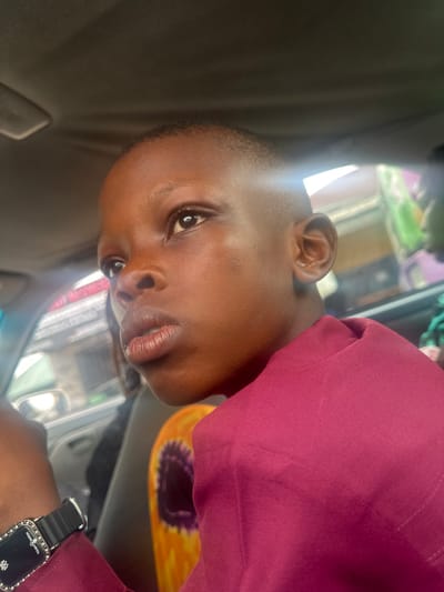 A young individual, appearing to be a child or pre-teen with dark skin and a short fade haircut, is depicted in a close-up, chest-up shot. The individual wears a maroon garment and a black rectangular digital watch with a silver-toned, stone-embellished bezel and "Tommy Watch" inscription on their left wrist. Their gaze is directed upwards and to the right, beyond the frame, with a neutral expression. The setting is inside a vehicle, evidenced by the gray car headliner visible at the top of the image and reflections on a window. Through the blurred vehicle window on the left, elements of an urban street scene are indistinctly visible, including part of another vehicle and what appears to be a building with faint red text signage. Behind and to the right of the subject, a vibrant orange and yellow patterned object with a distinct purple circular design is partially visible. The scene is located within a vehicle in the city of Elimgbu, Nigeria.