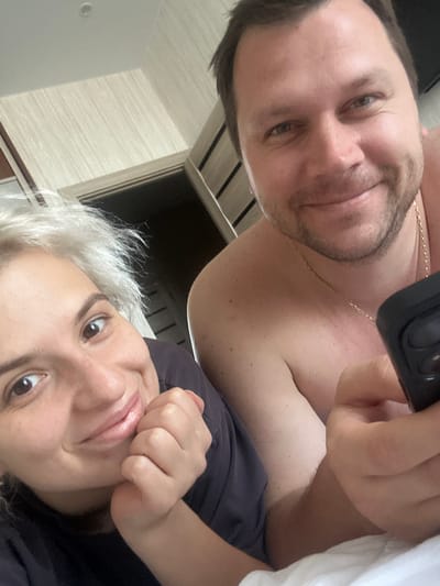 The image shows a selfie of a man and a woman in a bedroom. The woman is in the foreground, leaning on her hand and smiling. She has short, blonde hair and is wearing a dark shirt. The man is in the background, smiling and holding a phone. He has short, brown hair and is wearing a gold chain. The room appears to be a bedroom with light-colored walls and wooden furniture. The image was taken in the city of Lesnye Polyany, Russia.