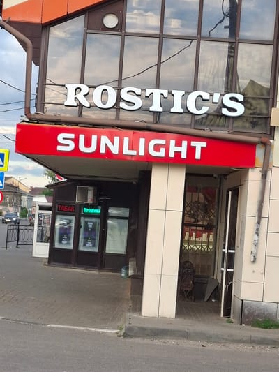 The image shows the storefront of a business with prominent signage. The primary subject is the building's facade, featuring a large red sign with white lettering that reads "SUNLIGHT" vertically. To the left of this sign, another vertical sign has white, three-dimensional letters spelling "ROSTIC'S". The storefront itself is made of glass panels and light-colored tiles.
The entrance to the store is visible, with a dark frame and glass doors. Inside, there are displays of products, including what appear to be cigarettes in a display case labeled "ТАБАК" (Tobacco). There are also electronic screens with green text and other illuminated advertisements. A metal grate or security gate is partially visible.
The scene appears to be on a street, with a paved sidewalk and road in the foreground. The time of day seems to be daytime, judging by the lighting. The weather is unclear, but the sky visible in the reflection on the glass is cloudy.
The location is identified as Меткомелино, Russia. The overall impression is that of a commercial establishment, likely a retail store selling various goods, including tobacco products. There are no people clearly visible in the image. The signage and architectural style suggest a commercial area.