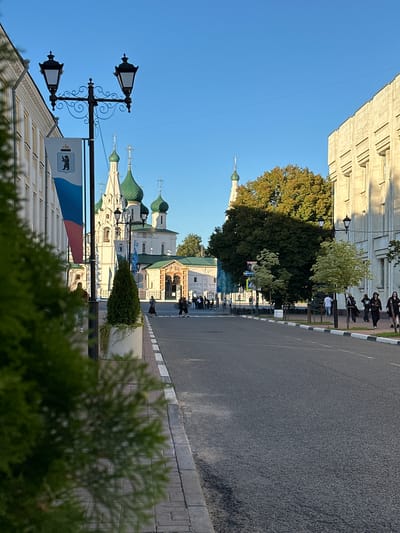 The image shows a street scene in Yazvitsevo, Russia, with a clear blue sky suggesting it is daytime, likely morning or afternoon. The perspective is unusual, rotated 90 degrees counter-clockwise, making the buildings appear to be on the left and the road on the right.
The foreground is dominated by the tops of green bushes and the side of a building with a white facade and patterned details. A street lamp with two lights is visible, along with a flagpole displaying a flag that appears to be the Russian tricolor with a coat of arms.
Further down the street, several domed buildings with green roofs, characteristic of Russian Orthodox churches, are prominent. These structures are white with decorative elements. A large, mature tree with dark green foliage stands near the churches, casting a shadow on the road.
On the right side of the image is a paved road, with a striped curb separating it from a brick sidewalk. Several people are walking or standing on the sidewalk, some appearing to be tourists or locals going about their day. The overall atmosphere seems peaceful and urban.
A sign with a depiction of a bear is visible on the flagpole. There is no discernible text in the image. The scene captures a slice of everyday life in a location with significant architectural and cultural features.