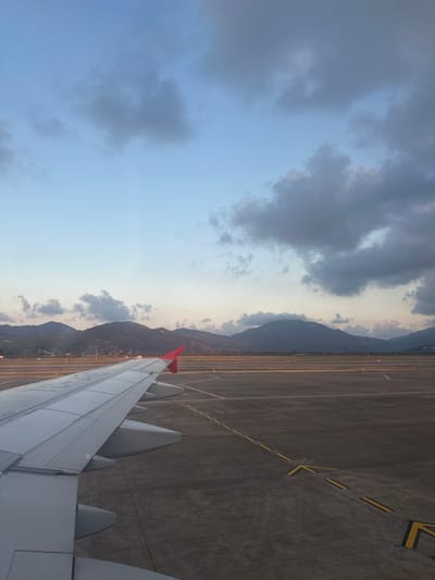 This image presents a detailed view from an aircraft window at Dalaman Airport (DLM) in Dalaman, Turkey, captured during either dawn or dusk. The left side of the frame features the light gray wing of an airplane, equipped with a prominent red wingtip (sharklet) on its outer edge. Faint lettering or a logo is discernible on the upper surface of the wing closer to the fuselage.

The foreground and midground to the right of the aircraft wing display dark gray asphalt or concrete airport ground, marked with distinct yellow dashed lines forming taxiway indicators and directional arrows. Additional white lines are visible further back on the airfield. Portions of the ground in the distance exhibit a warm, golden illumination.

Across the horizon, a range of dark, rugged mountains dominates the background, appearing sparsely vegetated. A faint line of human settlement or infrastructure, including subtle lights, is visible at the base of these mountains between the airfield and the peaks.

The sky transitions from a light blue with warm, soft hues near the mountain horizon to a darker, muted blue-gray in the upper portion. Scattered gray clouds, some with lighter edges catching distant light, are distributed throughout the upper half of the sky. No individuals are explicitly visible within the scene.
