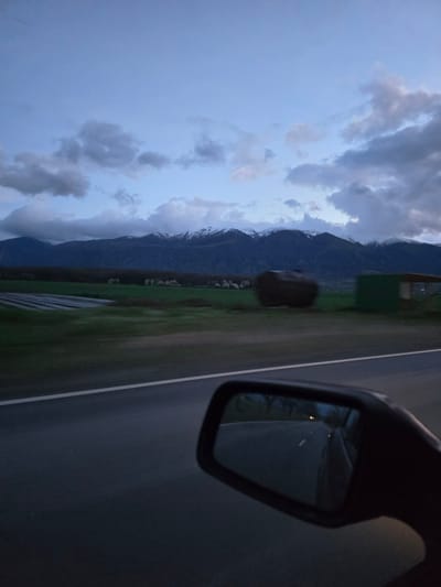 The image, taken from a moving vehicle, shows a view of mountains and a cloudy sky. The scene is likely taken in Marino Pole, Bulgaria, given the prompt's location information, though this cannot be confirmed visually. A side mirror of the car is visible in the upper right corner of the image, reflecting the road behind the car. To the left of the road is a green field with what appears to be a building or structure in the background. The mountains are visible in the distance, with low-hanging clouds obscuring their peaks.