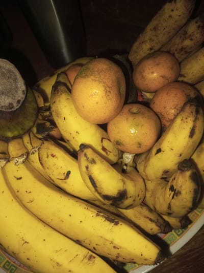 The image depicts a close-up, top-down view of a collection of fruit, likely situated indoors in Doramamidi, India. The primary subjects are numerous ripe yellow bananas and several oranges. The bananas, varying in size, exhibit yellow peels with scattered brown spots and occasional darker bruised areas. They are arranged in a pile, overlapping one another. Interspersed among the bananas are at least five oranges, characterized by their orange-yellow skin, some displaying green or brown blemishes. A fragment of a darker, possibly unripe or different fruit, with a fibrous or textured gray-brown end, is visible on the far left. The fruits are contained within a shallow, wide vessel, a portion of which is visible at the bottom and bottom-right, featuring a white rim with a geometric orange and green pattern. The background is dark and indistinct, suggesting an indoor setting with low, artificial overhead lighting creating shadows. No people or active interactions are present in the scene.