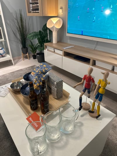 This image depicts an indoor scene, likely a living room or entertainment area. In the foreground, a white coffee table holds several items. These include two glass mugs, a red tag with "Novedad" printed on it, two brown glass bottles, two bags of chips, and two metal bowls on a wooden tray. Two wooden mannequin figures dressed as soccer players, one in red and one in yellow and blue, are positioned on the right side of the table, both on wooden stands. In the background, a large television screen displays a soccer game. To the left of the television is a wooden entertainment unit with white drawers. Further to the left, there are several potted plants, including a tall, slender plant and a large-leafed plant. A floor lamp with a white, textured shade is also visible. A part of a white shelf unit with a soccer ball is partially visible on the far left. The overall impression is a comfortable space set up for watching television and enjoying snacks and drinks. The mention of Siero, Spain, does not appear to be directly supported by visual evidence in the image, such as street signs or distinctive landmarks that would definitively place the location there.