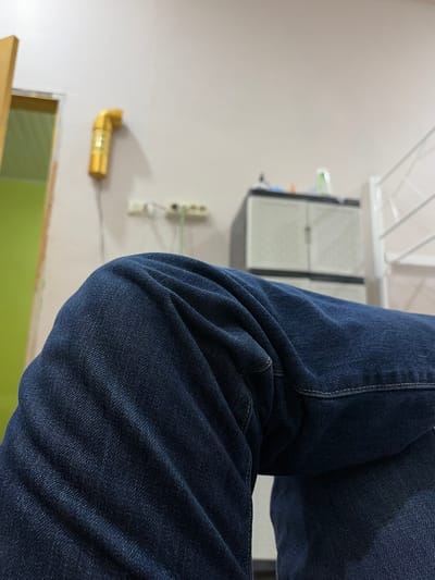 A low-angle, medium shot captures a person's bent knees, covered in dark blue denim jeans. The fabric of the jeans shows distinct creases and folds, particularly around the knee area. The background is lightly blurred, revealing a predominantly light-colored wall. To the left, a portion of a doorway with a green interior is visible, along with a wooden door frame. On the wall to the left, a bright yellow, curved object, possibly a piece of pipe or decoration, is mounted. Further back, a white electrical outlet with cords is visible. In the center of the background, a grey plastic cabinet with two doors stands against the wall. To the right, a white metal structure, likely a bed frame, is partially visible. The scene appears to be indoors, in a room that is sparsely furnished. The specific location in the city of Tupah, Indonesia, cannot be determined from the visual information provided.