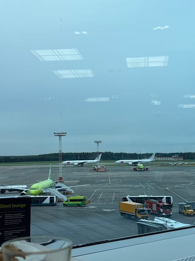 The image shows an outdoor airport scene, possibly in Kutuzovo, Russia, taken through a window with reflections from overhead lighting. In the foreground, a sign reads "Silent lounge" in English and Russian, with further information about flight status and boarding times. A blurry glass contains part of a drink.

The tarmac is visible, with several aircraft. A bright green airplane is parked, with a mobile staircase attached to its door, and a white bus and a small yellow vehicle nearby. Further away, two white airplanes with red and white markings are parked on the tarmac. Several support vehicles, including a red fire truck and a yellow towing vehicle, are also present. In the background, a dense line of trees forms the horizon, with some buildings and tall light poles scattered throughout the airport grounds. The sky is overcast and a pale blue. The reflections on the window include rectangular shapes and circular lights.
