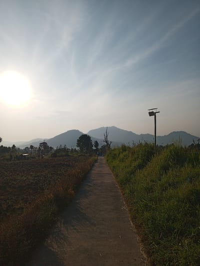 A paved pathway leads towards distant, hazy mountains under a pale blue sky with wispy clouds. The sun, partially obscured by atmospheric haze, is visible in the upper left quadrant of the image. To the left of the path, a field of dark, tilled soil with dry grass is depicted. To the right, a grassy verge is prominent, featuring a tall, rustic pole with a rectangular fixture on top, resembling a solar-powered lamp or birdhouse. In the mid-ground, scattered trees are visible, with a lone, bare-branched tree to the left of the path. Faintly, a person can be seen walking on the pathway in the distance. The overall environment suggests a rural or semi-rural setting, possibly on the outskirts of Yakkurugala, Sri Lanka, during daytime.