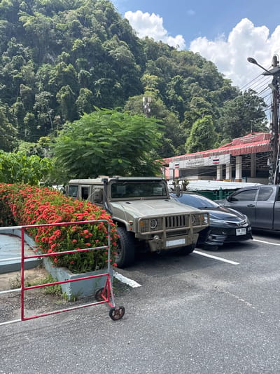 In Tambon Wang Prachan, Thailand, a desert tan HMMWV (High Mobility Multipurpose Wheeled Vehicle) is parked in an asphalt lot. The vehicle is positioned in front of a low wall overflowing with vibrant red Ixora flowers, also known as the flame-of-the-forest. Behind the HMMWV, a dark gray sedan and a pickup truck are also parked. Further in the background, a building with a red roof and white pillars is visible. The building has Thai signage that reads "Watthana Boundary Post" and "Bon Voyage". The backdrop is a lush, green mountainside covered in dense vegetation. A street light and utility pole are present on the right side of the image, with power lines stretching across the frame. A red and white metal barrier with wheels is situated in the foreground, partially obscuring the flower bed. The sky above is blue with scattered white clouds, and the overall lighting suggests it is daytime.