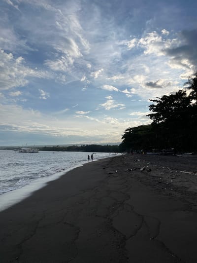 The image depicts a beach scene in Dauin, Philippines. The foreground shows dark, fine sand with some debris scattered on its surface, including plastic bottles, wood fragments, and what appear to be coconuts. The sand is textured with subtle patterns formed by the receding tide. To the left, the ocean waves gently roll onto the shore, creating a foamy white edge. The water is a deep grey-blue. In the mid-ground, a large white boat with multiple levels and flags is anchored offshore. Several people are visible in the water and walking along the shoreline in the distance.
The background features a lush, dark green treeline that runs along the coast. The trees are dense and appear to be tropical vegetation. The sky is a dramatic mix of blue and white, with scattered clouds that are illuminated by the sun, creating a sense of diffused light. The overall atmosphere suggests a late afternoon or early evening setting. The presence of the boat and people indicates recreational activity or local life on the beach.