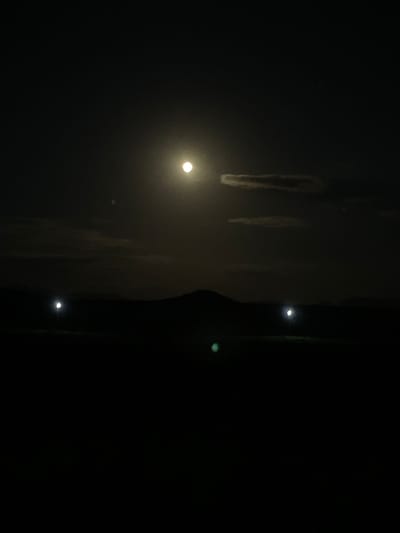 The image shows a night scene in Kara-Oi, Kyrgyzstan, dominated by a bright full moon in the upper center of the frame. The moon is surrounded by a soft halo of light, and a thin, horizontal cloud passes in front of it. Below the moon, the silhouette of hills or mountains is visible against the dark sky. In the middle ground, there are two distinct, bright light sources positioned at intervals, suggesting man-made lights, possibly from streetlights or buildings, on the horizon. The foreground is entirely dark, obscuring any details on the ground. A faint green light is visible in the lower center of the image. The overall impression is of a tranquil, nocturnal landscape illuminated by celestial and artificial lights.
