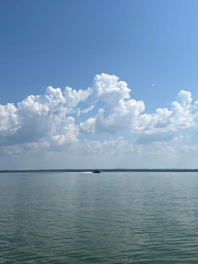 A wide shot captures a serene lake under a bright blue sky dotted with fluffy white clouds. A small boat with a dark hull is visible in the distance, creating a white wake as it cruises across the water. The horizon line is defined by a thin strip of land, possibly trees, separating the water from the sky. The water is calm with gentle ripples reflecting the light. The scene suggests a tranquil day in Bystrovka, Russia.