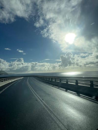 The image displays the Sea Cliff Bridge, situated within the Wollongong City Council area, Australia. The perspective is from an elevated vantage point on the bridge, looking forward along its curve. The two-lane asphalt road in the foreground appears dark and glistening, suggesting dampness, with two solid white center lines and embedded road markers. Concrete barriers topped with dark metal railings line both sides of the roadway. To the right, the Pacific Ocean extends to the horizon, reflecting a brilliant path of sunlight from the upper-right quadrant. A dark, distant coastal landmass or cliff is visible on the far left. The sky is bright blue, scattered with white cumulus clouds. The sun is intensely bright in the upper right, partially obscured by clouds, causing lens flare and strong illumination. No individuals, vehicles, or other distinct objects are present on the visible section of the bridge or in the immediate surroundings.