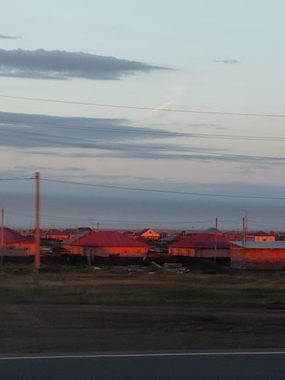 The image captures a residential area at dusk in Ayagöz, Kazakhstan. The sky is a pale blue with soft, elongated clouds tinged with pink and orange hues from the setting sun. Several houses with red-tiled roofs are visible, their walls illuminated by the warm, golden light. Electrical wires are strung across the sky, supported by poles. In the foreground, a strip of dark green grass lines a paved road. The overall scene is tranquil and suggests a rural or suburban setting.