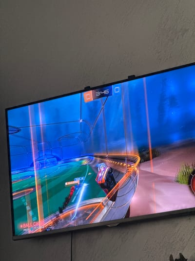 The image depicts a television screen displaying gameplay from the video game "Rocket League". The game shows two cars competing in a soccer-like match in an enclosed arena. The time remaining in the match is 3:46.
The location is not in Glojene, Bulgaria. There is no indication of the location.