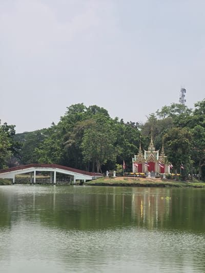This image depicts a serene waterside landscape in Phra Nakhon Si Ayutthaya City Municipality, Thailand. The right side of the frame is dominated by a calm, greenish-brown body of water, likely a lake or canal, displaying gentle ripples and subtle reflections.
Along the left bank, a lush expanse of green trees creates a dense, verdant backdrop. Nestled among the foliage are several small, ornate traditional Thai shrines or pavilions. These structures are characterized by their red walls, white bases, and multi-tiered roofs detailed with golden and green accents, reflecting typical Thai architectural styles. Small Thai flags, featuring red, white, and blue stripes, as well as some yellow and white flags, are visible near these shrines.
Further along the bank, a white bridge or walkway with reddish-brown railings extends, appearing to span or run alongside the water. Above the treeline in the upper-left, a modern communication tower equipped with multiple antennas stands out against the light, overcast sky, providing a contrast to the traditional and natural elements.
The soft, diffused lighting suggests an overcast day or a time of day without harsh sunlight. The scene is devoid of people, contributing to its tranquil and peaceful ambiance. No discernible text is visible in the image.