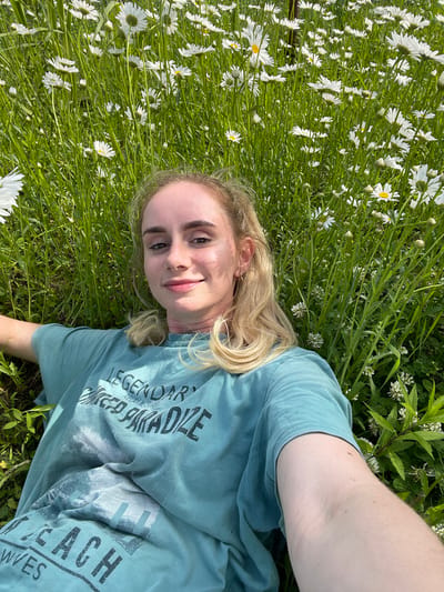 In Yasnoe, Russia, a young woman with blonde hair, wearing a teal t-shirt, lies on her back in a field of daisies and tall green grass, facing the camera. Her head is tilted slightly, and she is smiling. The t-shirt has a graphic print with text that reads "LEGENDARY," "RELAX & SURF," "PARADISE," and "BEACH WAVES." Her right arm is extended towards the camera, capturing a selfie. The background is filled with a dense meadow of white daisies and green foliage.