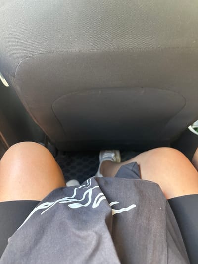 The image shows a person's legs from the knees down, sitting in the back seat of a car. The person is wearing a dark t-shirt with a white abstract design and black leggings. They are also wearing white sneakers. The background consists of a grey car seat and the car's floor mat. The image does not provide any information that can be used to determine the location in the city of Monastyr', Russia.
