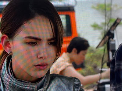 The scene is situated outdoors in Apshak, Uzbekistan. The primary subject is a young adult female featured in a close-up, occupying the left half of the frame. She has a fair complexion, dark brown hair pulled back from her face, and wears a small silver hoop earring in her left ear. Her eyes are dark, directed downwards and slightly to the right, conveying a serious or contemplative expression. She is wearing a silver or metallic grey jacket with a prominent, ridged collar.
In the blurred midground, to the right of the primary subject, a second individual, appearing male, is seen leaning forward. This individual has dark hair and wears a light-colored short-sleeved top, with a watch or bracelet visible on their left wrist. To the far right edge of the frame, the shoulder of a third person, wearing a dark plaid shirt, is partially visible. An orange-colored vehicle, resembling a utility truck, is partially visible in the left mid-background. Green foliage is discernible throughout the distant background, and a dark-colored object on a stand, possibly camera or lighting equipment, is visible in the right midground.