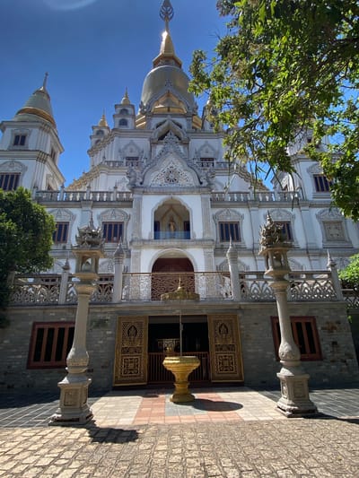 A detailed, slightly tilted, medium shot captures a portion of the Trúc Lâm Zen Monastery in Dĩ An, Vietnam. The monastery features a predominantly white facade, adorned with intricate ornamental detailing. The top of golden spires are visible against a clear blue sky. Parts of trees with green foliage are visible on either side of the building. The architectural style is characterized by layered structures, arched windows with dark blue frames, and elaborate stone carvings. Two ornate stone columns frame the entryway, leading to a golden gate with geometric floral patterns. The ground is paved with light-colored brick.