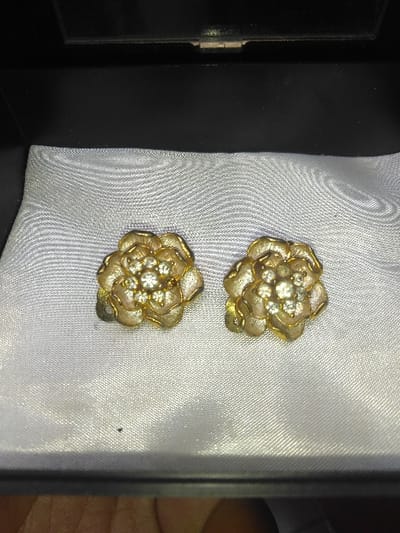 The image displays two identical flower-shaped earrings, each crafted from gold-toned metal with a central cluster of multiple small, clear, faceted stones. They are positioned side-by-side on a padded, off-white satin-like fabric liner within an open, black rectangular box. The interior upper edge of the box is visible, featuring a dark, slightly reflective surface. A segment of a human hand, exhibiting light brown skin tones, is partially visible at the bottom-left corner of the frame. The specific location within the city of Liège, Belgium, cannot be determined from the visual information provided in this image.