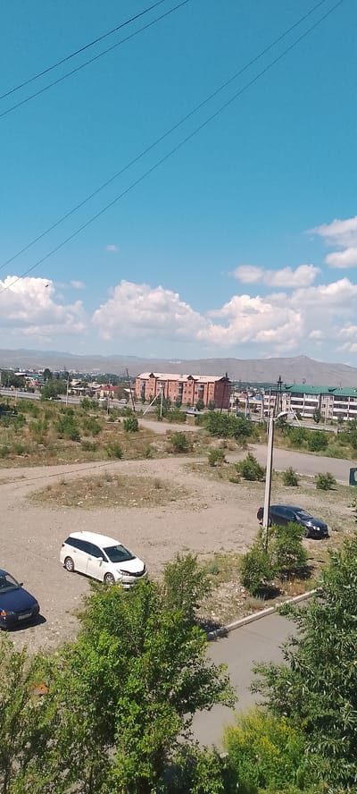 This is a medium-shot, eye-level, outdoor, vertical photograph taken on a sunny day in the city of Kyzyl, Russia. The sky is bright blue with scattered white clouds. Several power lines are visible in the upper portion of the frame.

In the mid-ground, there is a large, multi-story brick building with a light-colored roof and balconies. To the right of this building are several other buildings, some with green roofs. Power poles and lines are present throughout this area. The terrain is mostly dry and dusty with sparse vegetation.

In the foreground, on a dirt and gravel surface, are three cars: a dark blue sedan on the left, a white station wagon in the center, and a dark gray sedan to the right. The white station wagon appears to be parked. Lush green trees are visible in the immediate foreground, obscuring the lower portion of the scene. The background shows rolling hills under a bright sky.