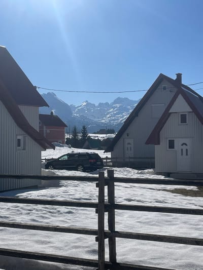 The image depicts a winter or early spring scene in Palež, Montenegro, under a clear, bright blue sky with direct sunlight. In the immediate foreground, a weathered, dark brown split-rail wooden fence with multiple horizontal rails extends across the lower portion of the frame. Beyond the fence, the ground is covered in uneven patches of melting snow, revealing some bare earth.
The midground features three distinct houses with white vertical siding and steep reddish-brown gable roofs. On the far left, a two-story house has one visible window on its left side. To the right, a larger two-story house exhibits two white doors on its front facade, suggesting multiple entrances, and a single small window above the right door. A chimney is visible on its roof. Partially obscured on the far right is a third, smaller white house with a similar roof style and one visible white door.
A dark-colored, possibly black, station wagon is parked between the left and center-right houses, partially embedded in the snow, facing away from the viewer. Further in the midground, a red-colored house is visible between the foreground structures, nestled among snow-covered terrain and sparse evergreen trees. A thin black utility line is visible stretching horizontally across the upper midground, partially obscuring the distant mountains.
The background is dominated by a majestic range of rugged, snow-capped mountains under the clear sky. The mountains exhibit prominent white snow coverage interspersed with darker rock formations. The sun's glare is visible as a bright lens flare in the upper left quadrant of the sky. No individuals or animals are present in the scene, and no specific actions or interactions are depicted.