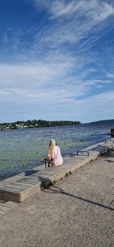 A woman with long blonde hair, wearing a pink top and a leopard print bag, is bent over on a wooden boardwalk beside a lake. The lake is covered in lily pads and appears to be a natural body of water. The boardwalk runs alongside the lake, and beyond that is a gravel path with some grass growing along the edge. In the background, a tree-lined shore is visible, with houses and buildings scattered among the trees, suggesting a populated area. The sky is blue with wispy white clouds. The sun is shining, indicating it is daytime and the weather is likely warm. The overall scene suggests a peaceful, natural setting, possibly a recreational area or a lakeside town. The woman's posture suggests she might be looking at something in the water or adjusting something on the boardwalk. The image appears to be taken from a slightly elevated angle. There is no visible text in the image.
