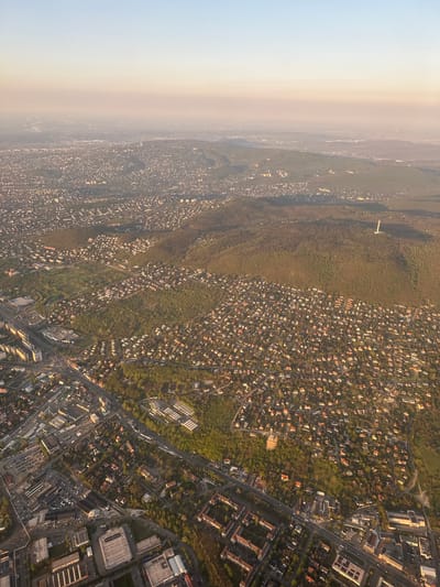 The image is an aerial view of a suburban area, possibly in the vicinity of Vecsés, Hungary. The photograph shows a dense concentration of residential buildings, a mix of houses with orange rooftops intermingled with greenery. There is also a clear distinction between densely populated residential areas and larger institutional or commercial buildings with rectangular rooftops, especially in the lower-right section of the photo. There appears to be a forest or green space, and a horizon with what looks like the edge of the city in a hazy atmosphere. The orientation of the image suggests that the photograph was taken from an airplane.