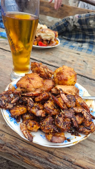 A close-up, eye-level shot displays a platter of cooked shrimp and chicken pieces, coated in a dark, glossy sauce, resting on a white plate with blue floral trim. The plate is positioned on a rustic wooden table. To the left, a tall glass filled with golden beer stands prominently, its condensation suggesting it is cold. In the background, another plate holds what appears to be small bread rolls and pieces of red and yellow bell peppers, partially obscured. A person's hand is visible in the upper right corner, reaching towards the food. A blue and white checkered blanket is draped behind the food, also partially obscuring a wooden bench or surface. The overall scene suggests an outdoor dining setting, possibly a picnic or casual meal. The specific location in Панфилово, Kazakhstan, is not visually indicated within the image.