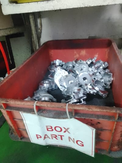 The image shows a red plastic bin filled with small, metallic parts. The bin is placed on a green surface, possibly a factory floor. The metallic parts appear to be machine components, possibly for an automotive or industrial application, given their intricate designs with holes and flanges. They are shiny and silver, suggesting they are made of aluminum or a similar metal.

Attached to the front of the bin is a white label with red lettering. The visible text on the label reads "BOX PART NG". This likely indicates that the contents are a box of parts, and "NG" could be an abbreviation for "No Good" or "Non-conforming", suggesting these parts are either scrap, awaiting inspection, or have failed a quality check.

The setting appears to be an industrial or manufacturing environment, as suggested by the bin, the parts, and the overall utilitarian appearance of the surroundings. The lighting is artificial, casting some shadows and highlighting the metallic sheen of the parts. There are no people visible in the image. The image does not provide any specific time of day or weather cues, but the overall impression is one of an active industrial workspace. The location context of Ciantra, Indonesia, further supports the industrial setting.