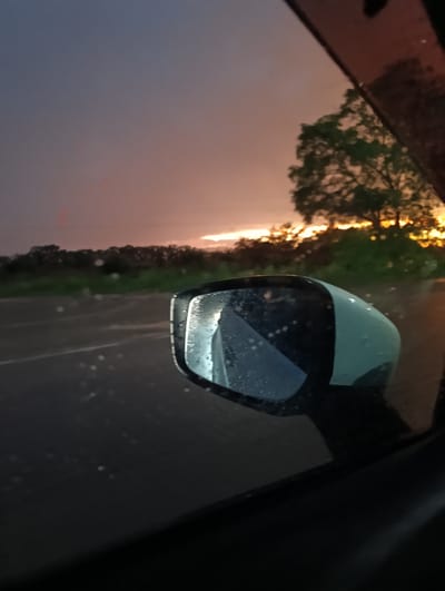 This media file shows a view from inside a car, looking out the side window towards a side mirror. The side mirror reflects the sky, which is a dusky purple and orange, indicating it is either sunrise or sunset. Raindrops are visible on the mirror, suggesting it is raining or has recently rained.

In the background, there is a road with white lines, suggesting it is a highway or main road. Beyond the road, there is a line of dark trees and green foliage. A large tree with green leaves is prominent on the right side of the image. The overall scene evokes a sense of travel during inclement weather with a beautiful sky. The emotional tone is calm and possibly reflective, due to the quietness of the scene and the muted colors of the sky.

The image is a photograph taken in Ptichnik, Russia. There is no visible text in the image.