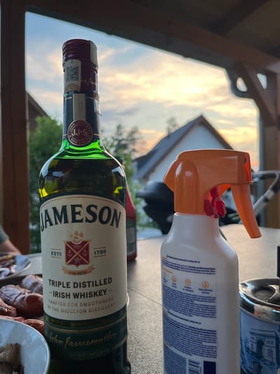 This is a close-up, eye-level, outdoor shot taken during sunset, focusing on a bottle of Jameson Irish Whiskey and a bottle of sunscreen. The Jameson bottle, which is green glass with a prominent label, is positioned on the left, slightly angled towards the camera. The label displays "JAMESON," "TRIPLE DISTILLED," "IRISH WHISKEY," and "CRAFTED FOR SMOOTHNESS AT THE MIDLETON DISTILLERY," along with the brand's crest and establishment date of 1780. To the right of the whiskey bottle is a white plastic bottle with an orange spray nozzle, identified as a sunscreen product with SPF 30. The background shows a blurred view of trees, houses with dark roofs, and a twilight sky with orange and purple clouds. A table is visible in the foreground, with hints of food, possibly sausages, on a plate to the left, and a metallic object, possibly an ashtray, to the right. The overall setting appears to be a patio or balcony in a residential area, possibly during a gathering or meal. The specific location in the city of Milná, Czechia, cannot be confirmed from the visual information provided.