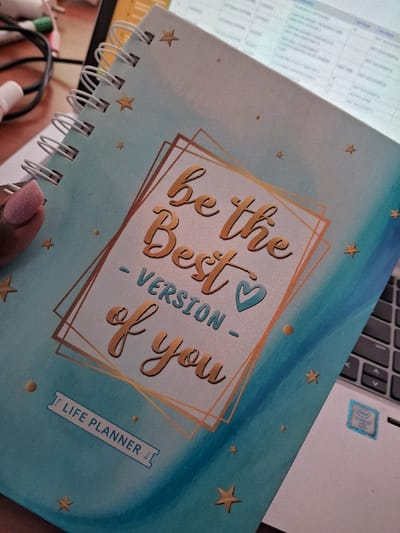 The scene depicts an indoor setting in Muthambi ward, Kenya, focusing on a desk area. A light blue and teal gradient spiral-bound "LIFE PLANNER" notebook is centrally positioned, held by a human left hand with a light pink, possibly glittered, fingernail. The planner's cover features gold foil text: "be the Best VERSION of you," with "VERSION" in blue, accompanied by a small blue heart icon and enclosed within a geometric gold frame. Gold stars and dots are scattered across the cover. White wire spiral binding is visible on the left edge of the planner. A dark-colored laptop is partially visible in the background and to the right, showing parts of its keyboard with dark gray keys and a white sticker bearing an Intel Core i7 logo. The laptop screen, out of focus, displays tabular data in light blue and white cells. Additional out-of-focus white objects and black cables are visible in the upper left background.