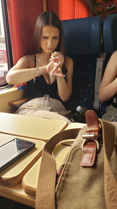 A young woman with long brown hair sits on a train, looking at her watch with a concerned expression. She is wearing a black top and light-colored pants, adorned with several bracelets. A mobile phone is visible on the table in front of her. The train interior features blue seats and orange curtains. The image does not provide any information about the city of Púchov, Slovakia.
