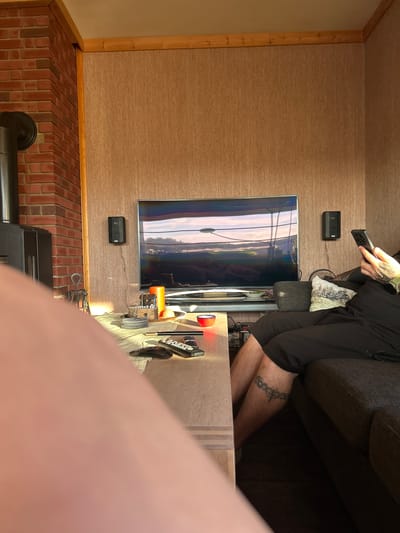 This is a medium shot, indoors, of a living room space, with the foreground blurred by a person's arm and leg. The background shows a television with a landscape scene, a brick fireplace with a wood-burning stove pipe, and a person sitting on a couch using a smartphone. The walls are covered with a textured, light brown wallpaper, and there is a wooden trim along the ceiling. The floor appears to be carpeted. There are speakers mounted on the wall next to the television. On a light-colored wooden table in front of the couch, there are several remotes, a small decorative house, an orange candle, and a small red bowl. The person on the couch is wearing black shorts and has a tattoo on their left thigh. The lighting suggests it is daytime, with sunlight streaming in from the right. The specific location in Kongsberg, Norway is not discernible from the visual information in the image.