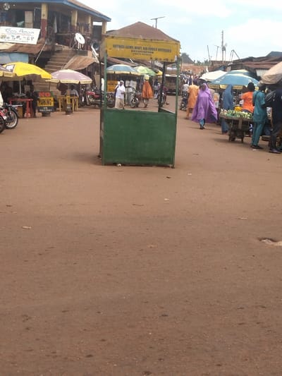 The image shows a street scene in Lokoja, Nigeria, located at coordinates 7.404 latitude and 7.632 longitude.  A green and yellow kiosk with the inscription "Retired Army Navy Air Force Officers Association Kogi Branch" is prominent in the foreground. Several people are present, some walking on the dusty street, others are sitting on chairs or standing near the kiosk. A motorcycle is parked on the left side of the image. A "No Parking" sign is visible on the left side of the street, indicating the street is a designated parking zone.  The street is unpaved, dusty, and appears to be in a residential or commercial area. There are several stalls and shops in the background, suggesting the location is a marketplace. The sky is overcast with some cloud cover.  The scene depicts a typical everyday street scene in Lokoja, Nigeria.