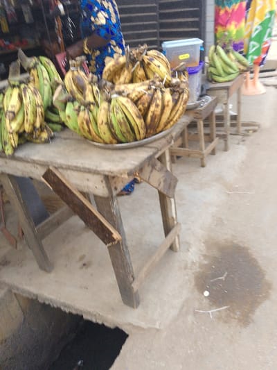 This image depicts an outdoor market scene, likely in Ijaiye, Nigeria, showcasing a stall primarily selling bananas.
The central focus is a large quantity of bananas displayed on two rustic wooden tables or stands. On the main wooden table in the foreground, a substantial collection of bananas, varying from green with brown streaks to more yellow-brown, fills a round, silver-colored metal tray. Another smaller bunch of greener bananas is positioned to the left of the tray on the same table. Further back on the right, a smaller wooden stand holds more bananas, and a translucent light blue plastic container with a yellow label sits beside them. A third, smaller wooden stool is visible between the two main stands.
A person is partially visible behind the main display, wearing a blue garment with yellow patterns, possibly a traditional wrapper or dress. Their left arm is visible, adorned with a gold-colored bangle, suggesting they are attending the stall.
The setting appears to be a street or outdoor market. The ground is paved concrete, with a noticeable dark, wet patch and some small debris including twigs and a white pebble. In the bottom-left corner, a dark, open drain or ditch is visible. In the background, to the right, colorful patterned fabrics are draped, possibly for sale or as part of another stall's display. Behind the main stall, a dark, slatted or corrugated wall can be seen.
The lighting suggests it is daytime. No specific emotions are discernible from the partial view of the person, and no legible text is visible on any objects, though the plastic container has a label. The overall impression is a bustling, informal market environment.