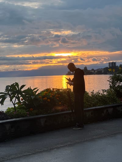 The image displays a solitary male figure on a paved promenade adjacent to Lake Geneva in Montreux, Switzerland, at sunset. The man, depicted in profile, is silhouetted against the horizon, where the setting sun casts a strong orange and yellow glow across the sky, partially obscured by horizontal cloud formations. He is observed holding and viewing a smartphone with both hands, looking down at the device. A closed black umbrella is held vertically in his left hand. The surface of Lake Geneva reflects the vibrant sunset colors, appearing bright orange and yellow across the midground. In the immediate foreground to the left, a low, dark stone wall supports a cultivated flower bed containing orange marigold-like flowers and various green foliage. Distant landmasses and structures, including multi-story buildings, are faintly visible on the far shore across the lake. The scene indicates an outdoor, public lakeside environment, consistent with a pedestrian walkway.
