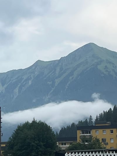 The image depicts a mountainous landscape in Böckstein, Austria, under an overcast sky. In the foreground, a large, dense green tree dominates the lower left portion of the frame, with glimpses of buildings and structures partially obscured behind it and to its right. These buildings appear to be of modern construction, featuring yellow and dark-colored facades with visible windows and balconies. A dense forest of evergreen trees lines the mid-ground, ascending the slopes of the mountains. A thick layer of white clouds or fog blankets the lower sections of the mountains, creating a soft, diffused effect and obscuring the base of the peaks. The mountains themselves are rugged and covered in lush green vegetation, with visible textures and shadows suggesting steep inclines. The overall atmosphere is subdued, consistent with a cloudy or misty day. There are no people visible in the frame.