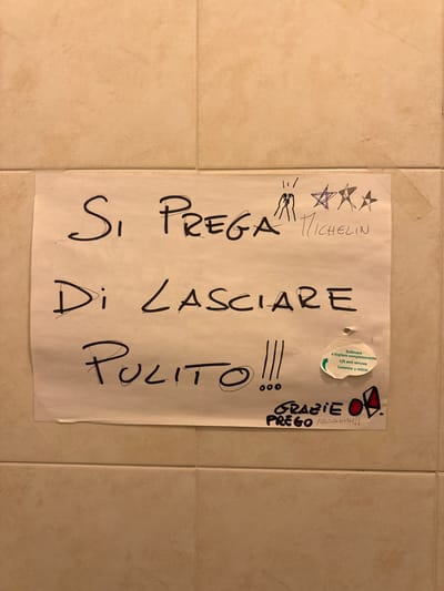 The image shows a handwritten note on a piece of paper attached to a tiled wall, likely in a public restroom in Fontevivo, Italy. The note, written in Italian, reads "SI PREGA DI LASCIARE PULITO !!!" which translates to "PLEASE LEAVE CLEAN !!!". Above this text, there are some drawings, including what appears to be hands, stars, and the word "MICHELIN". Below the main message, there is a sticker in English, Italian, and Spanish with instructions to "Sollevare e togliere completamente / Lift and remove / Levantar y retirar" accompanied by a green arrow. At the bottom of the note, there is another handwritten message in Italian: "GRAZIE PREGO NAAAHH !!!". The tiled wall appears to be a light beige color with grout lines. The lighting suggests an indoor setting.
