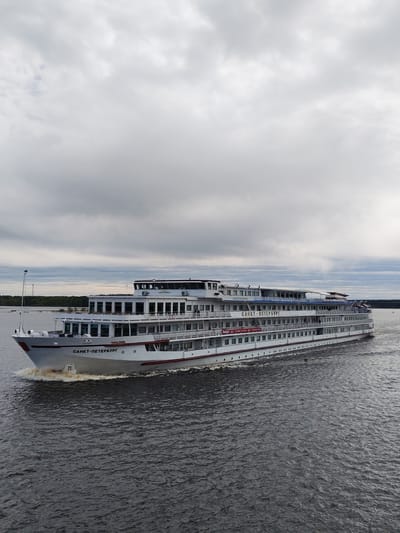 The image captures a large, multi-decked river cruise ship named "Sankt-Peterburg" moving on a body of water, possibly a river or lake. The ship is white with red trim, and the name is written in Cyrillic on its bow and side. The sky is overcast with grey clouds. In the background, a strip of land is visible, suggesting a shoreline. The water is dark and choppy, indicating some wind or current. The setting could be the city of Myshkin, Russia, as the ship's name and the environment are consistent with Russian river cruises. No people are clearly visible in the image. The focus is on the ship and its movement through the water.